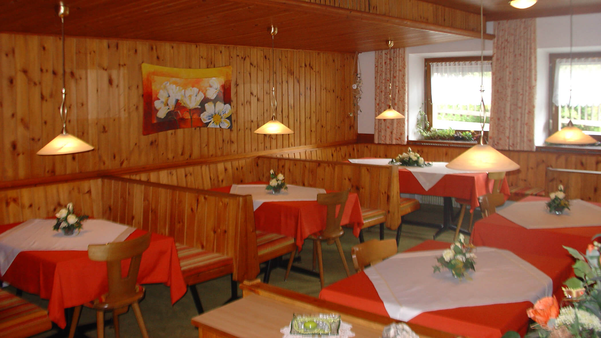 Ein gemütlicher Frühstücksraum mit Holzwänden, roten Tischtüchern, Blumenschmuck und Hängelampen. - Garni Tschurtschenthaler