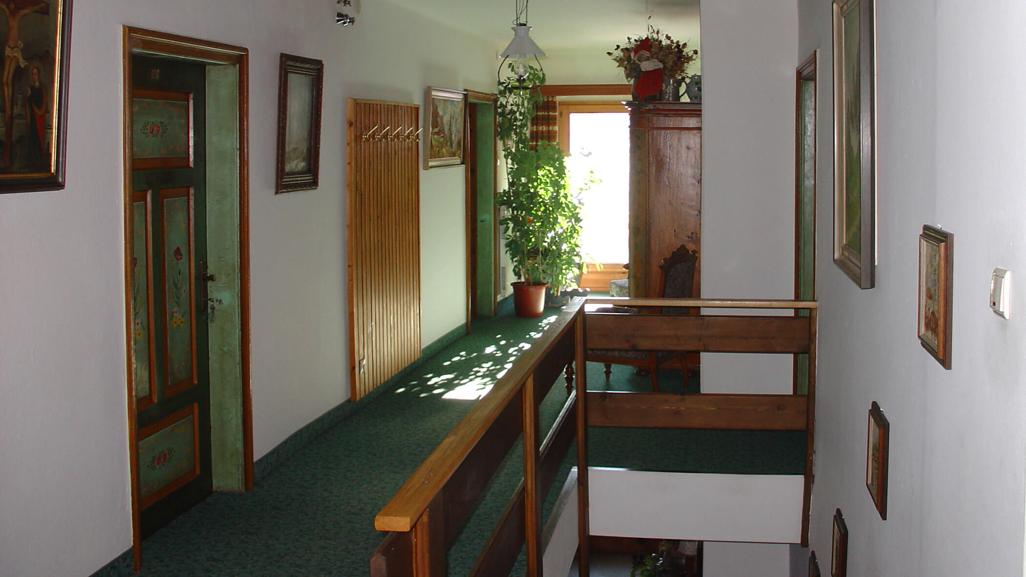 Ein sonnendurchfluteter Flur mit grünem Teppich, Holztüren, Topfpflanzen und gerahmten Bildern an weißen Wänden. - Garni Tschurtschenthaler