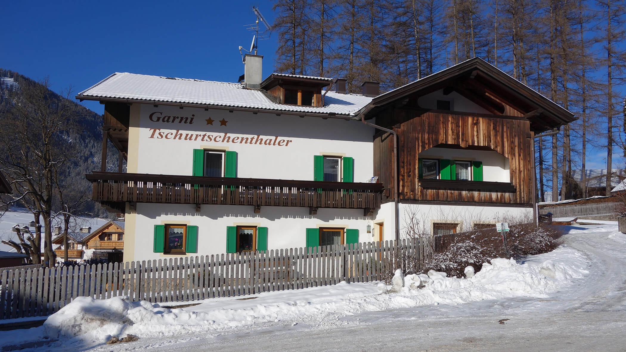 Weißes und hölzernes alpines Gästehaus mit grünen Fensterläden, verschneitem Boden und Pinienbäumen unter einem klaren blauen Himmel. - Garni Tschurtschenthaler