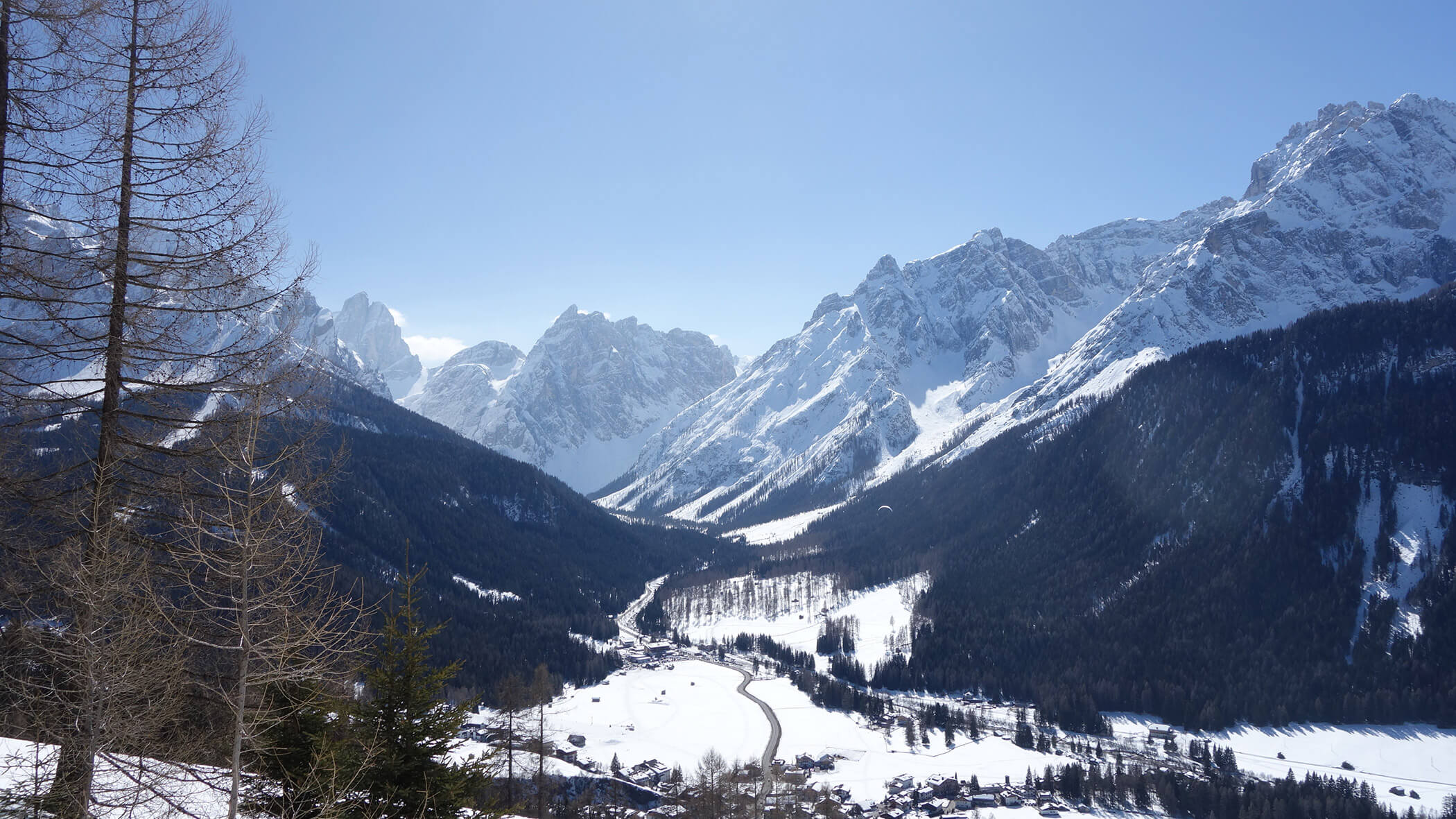 Schneebedeckte Berge mit einem Tal, einer kurvenreichen Straße und vereinzelten Bäumen unter einem klaren blauen Himmel. - Garni Tschurtschenthaler