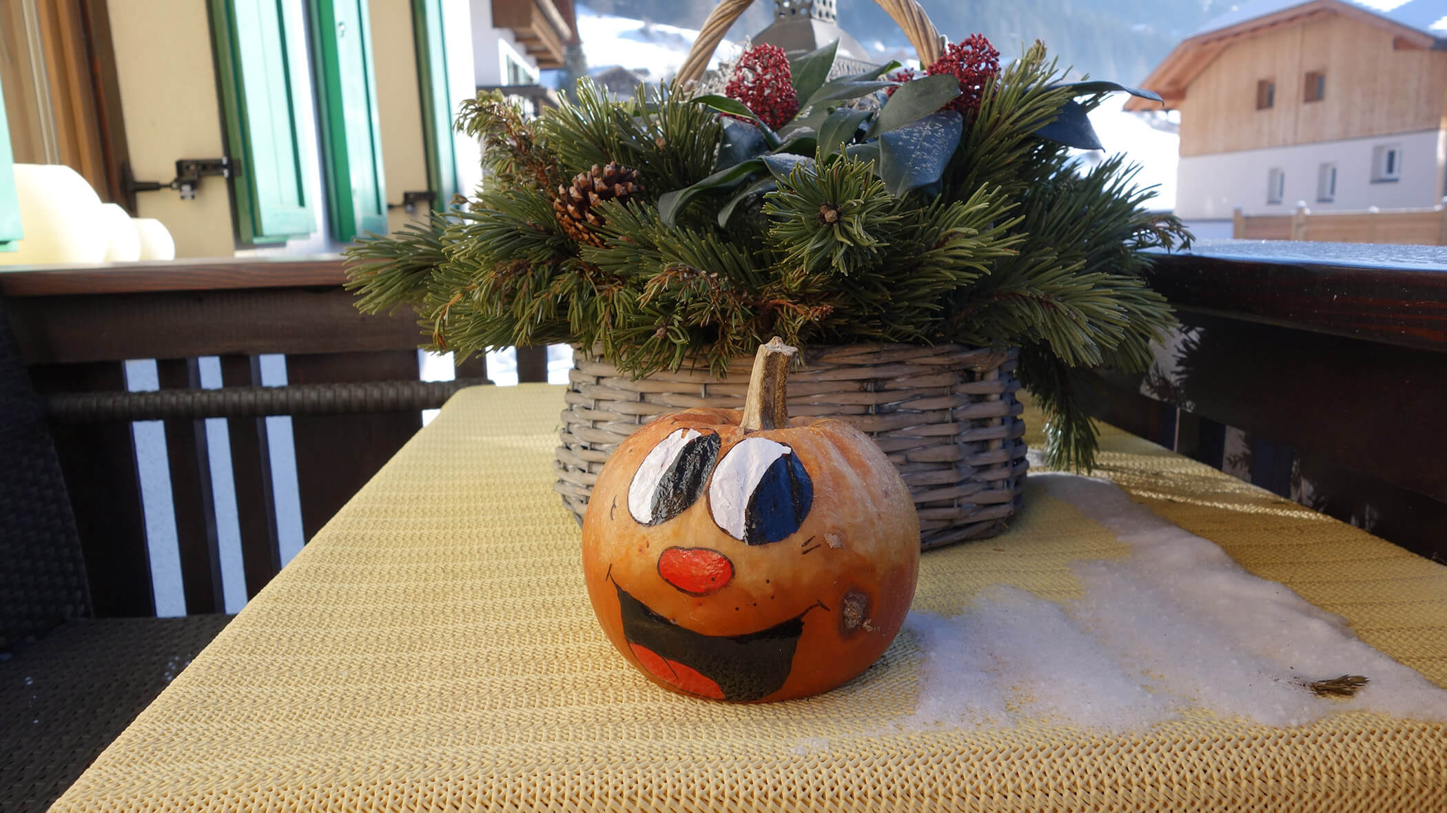 Ein kleiner Kürbis mit einem aufgemalten Gesicht sitzt auf einer gelben Tischdecke neben einem Korb mit immergrünen Zweigen. - Garni Tschurtschenthaler