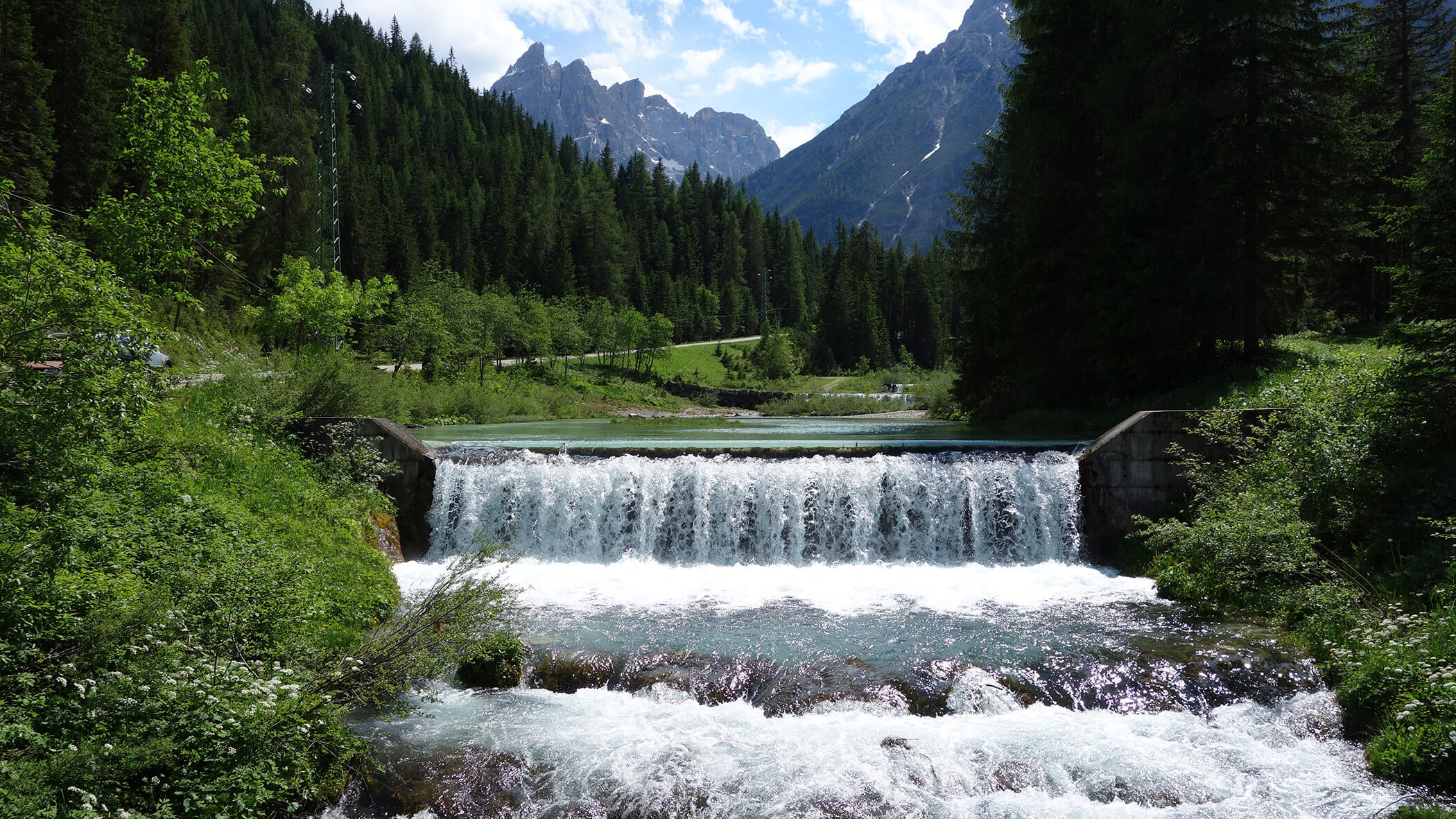 Ein kleiner Wasserfall fließt durch einen Wald mit Bergen im Hintergrund unter einem teilweise bewölkten Himmel. - Garni Tschurtschenthaler