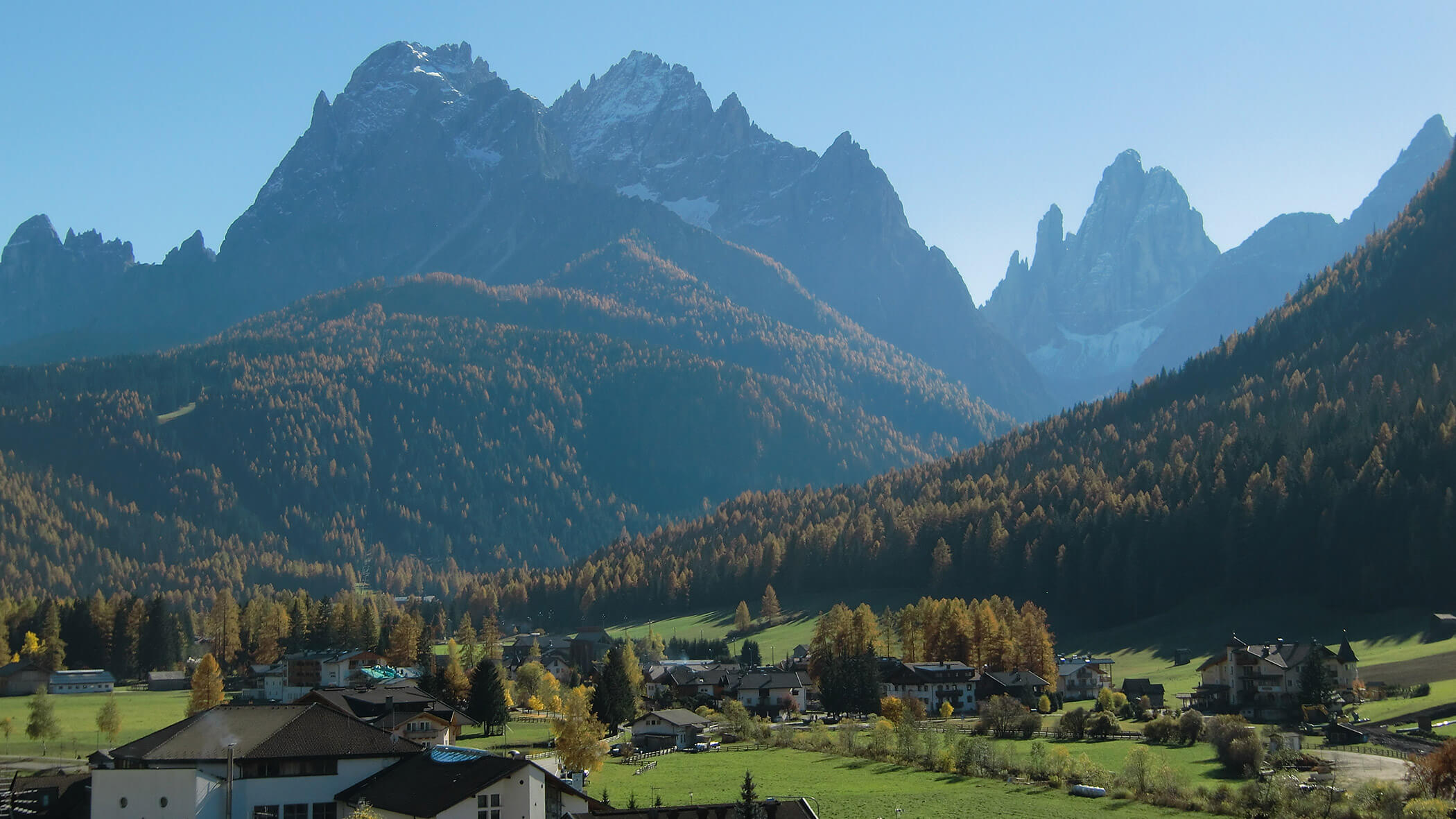 Ein kleines Dorf liegt in einem grünen Tal, umgeben von bewaldeten Hügeln und hohen, schroffen Bergen. - Garni Tschurtschenthaler