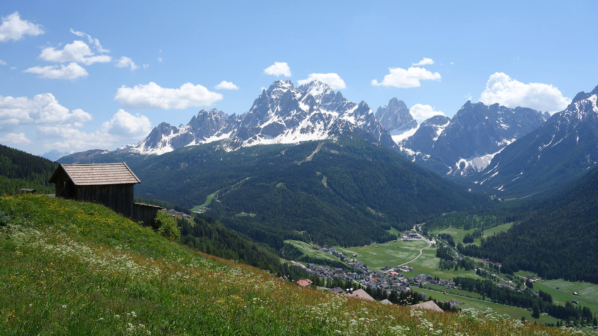 Alpinwiese mit Holzhütte, Blick auf ein Tal und schneebedeckte Berggipfel unter blauem Himmel. - Garni Tschurtschenthaler