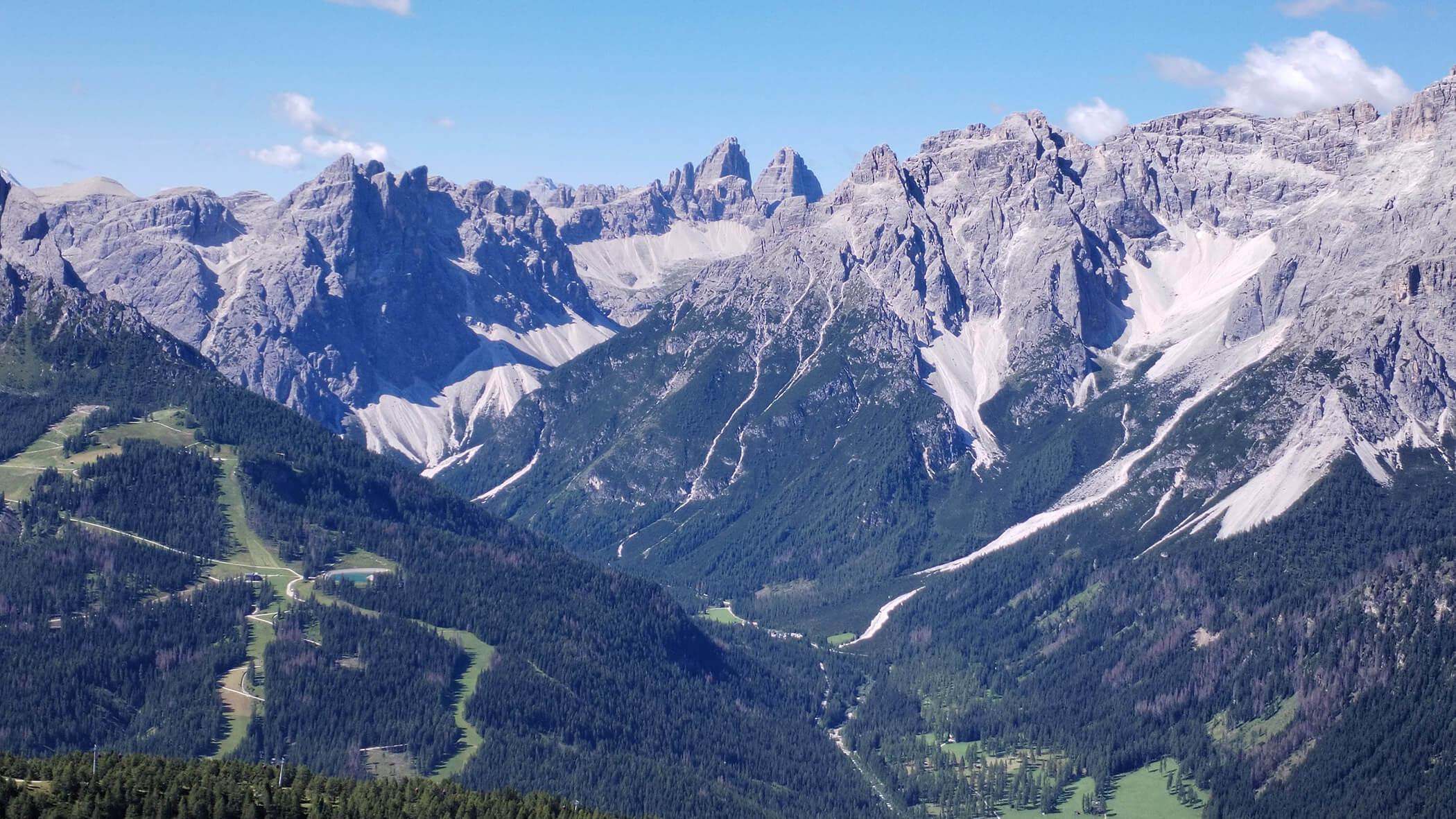 Zackige Berggipfel erheben sich über grün bewaldete Täler unter einem klaren blauen Himmel. - Garni Tschurtschenthaler