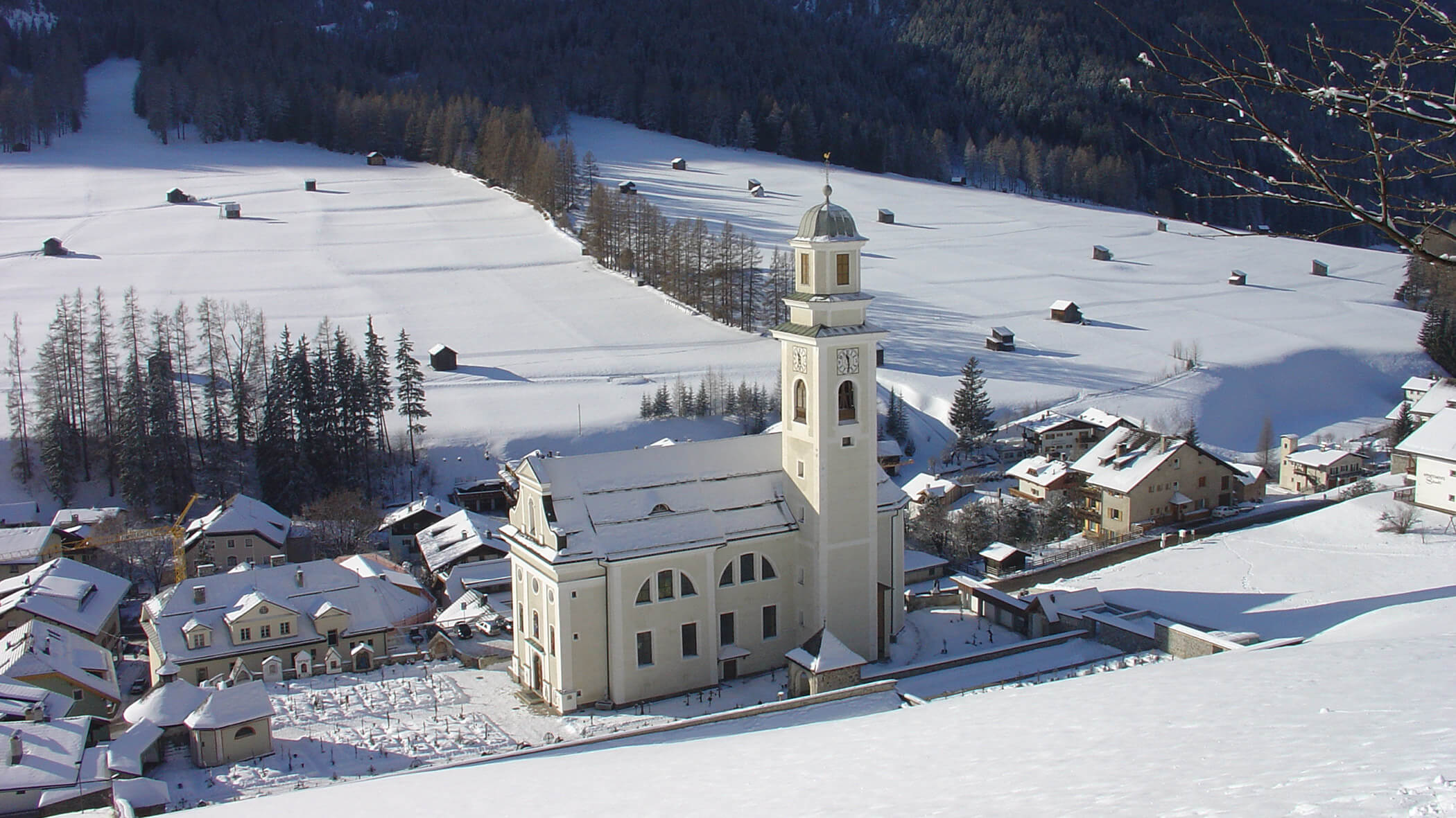 Eine weiße Kirche mit einem Glockenturm steht in einem verschneiten Dorf, das von Bäumen und schneebedeckten Feldern umgeben ist. - Garni Tschurtschenthaler