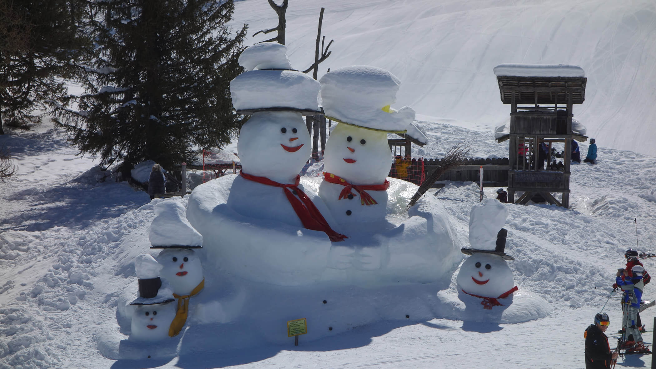 Fünf Schneemänner mit Mützen und Schals stehen zusammen in einem verschneiten Außenbereich in der Nähe von Bäumen und Holzkonstruktionen. - Garni Tschurtschenthaler