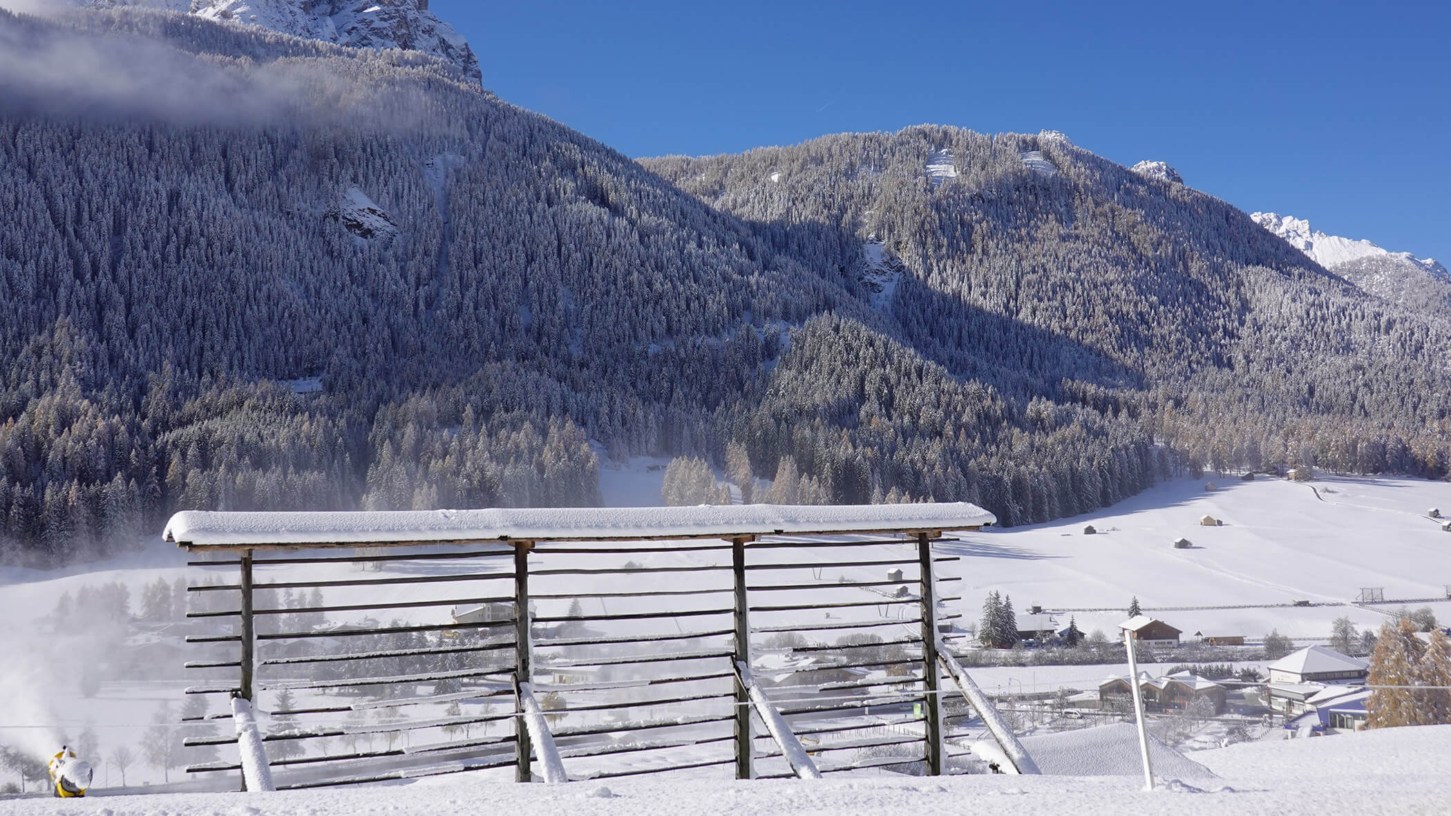 Schneebedecktes Holzgestell vor einem verschneiten Tal mit bewaldeten Bergen unter einem klaren blauen Himmel. - Garni Tschurtschenthaler