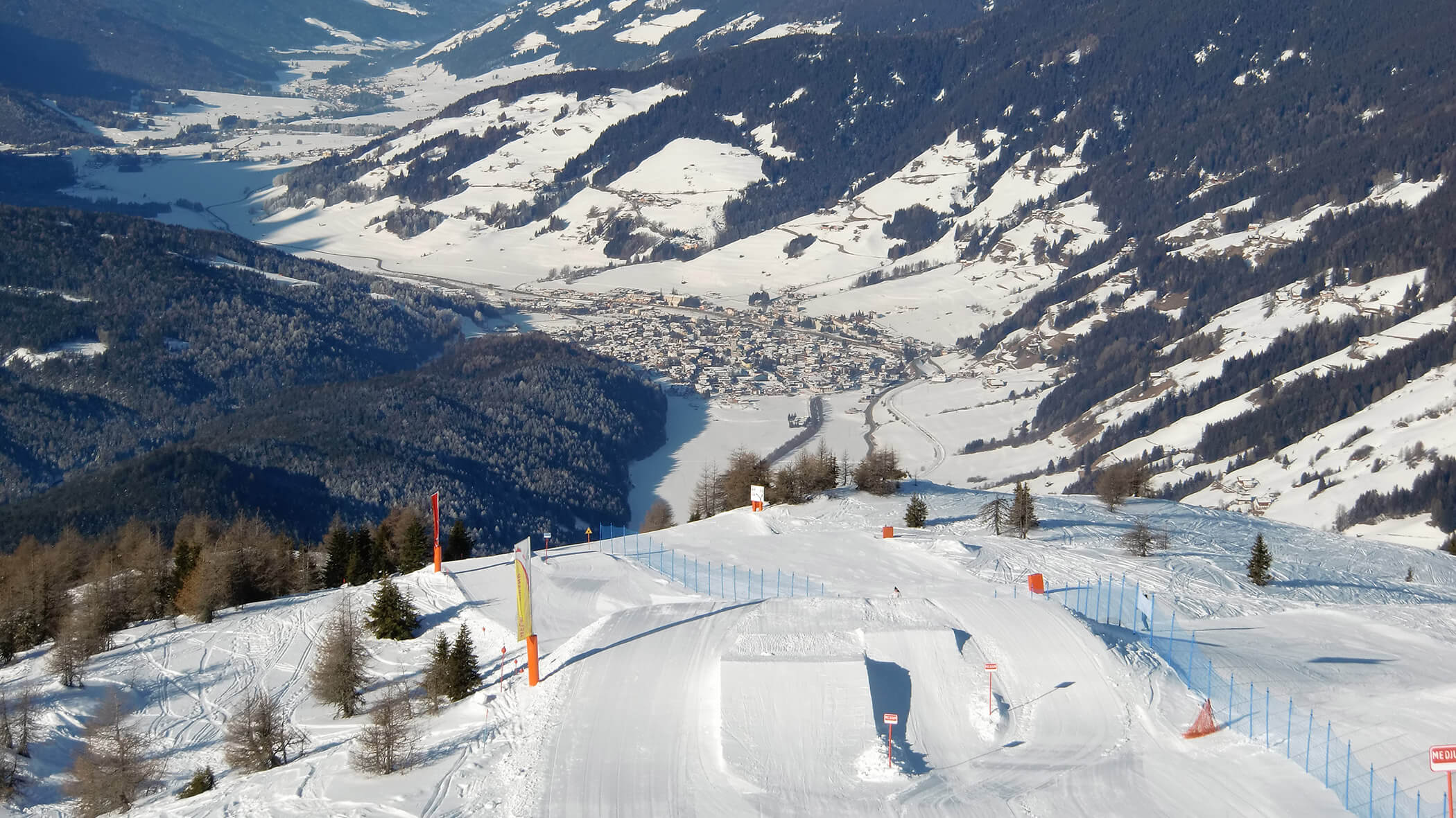 Eine verschneite Skipiste mit Schanzen überblickt ein Tal und eine Stadt, die von schneebedeckten Bergen umgeben ist. - Garni Tschurtschenthaler