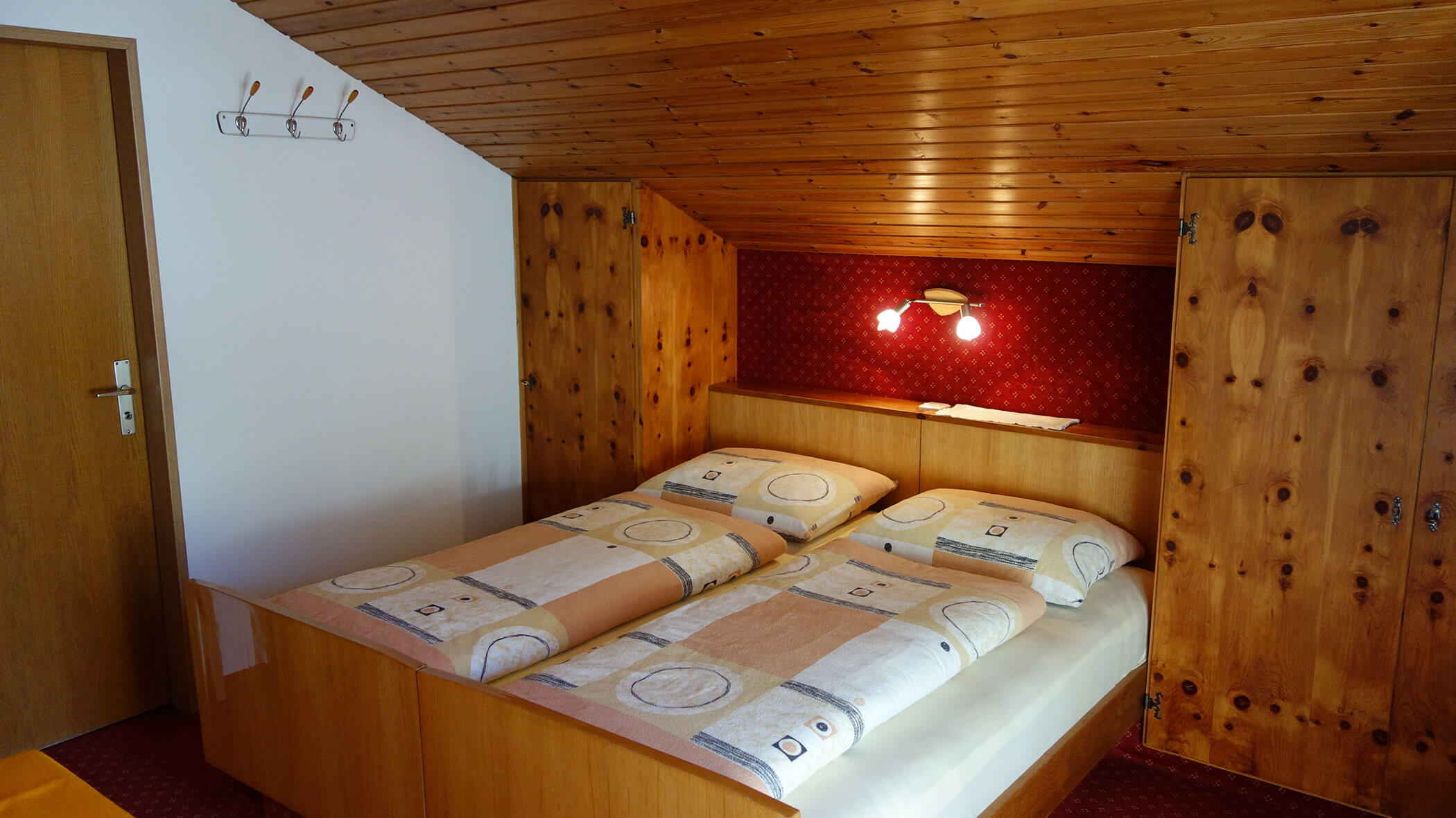 Gemütliches Schlafzimmer mit zwei Einzelbetten, Holzmöbeln, schräger Decke und einer Wandlampe über dem Kopfteil. - Garni Tschurtschenthaler