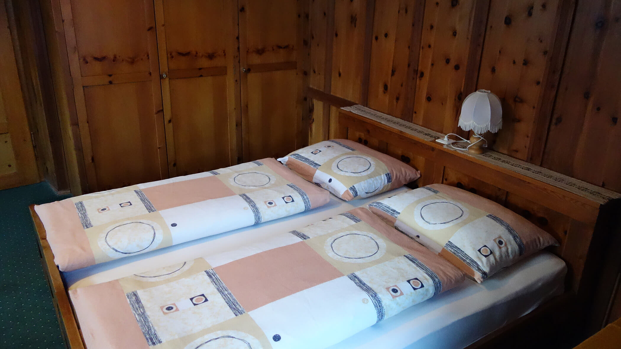 Doppelbett mit geometrisch gemustertem Bettzeug, Kopfteil aus Holz und einer kleinen Lampe auf einem schmalen Regal. - Garni Tschurtschenthaler