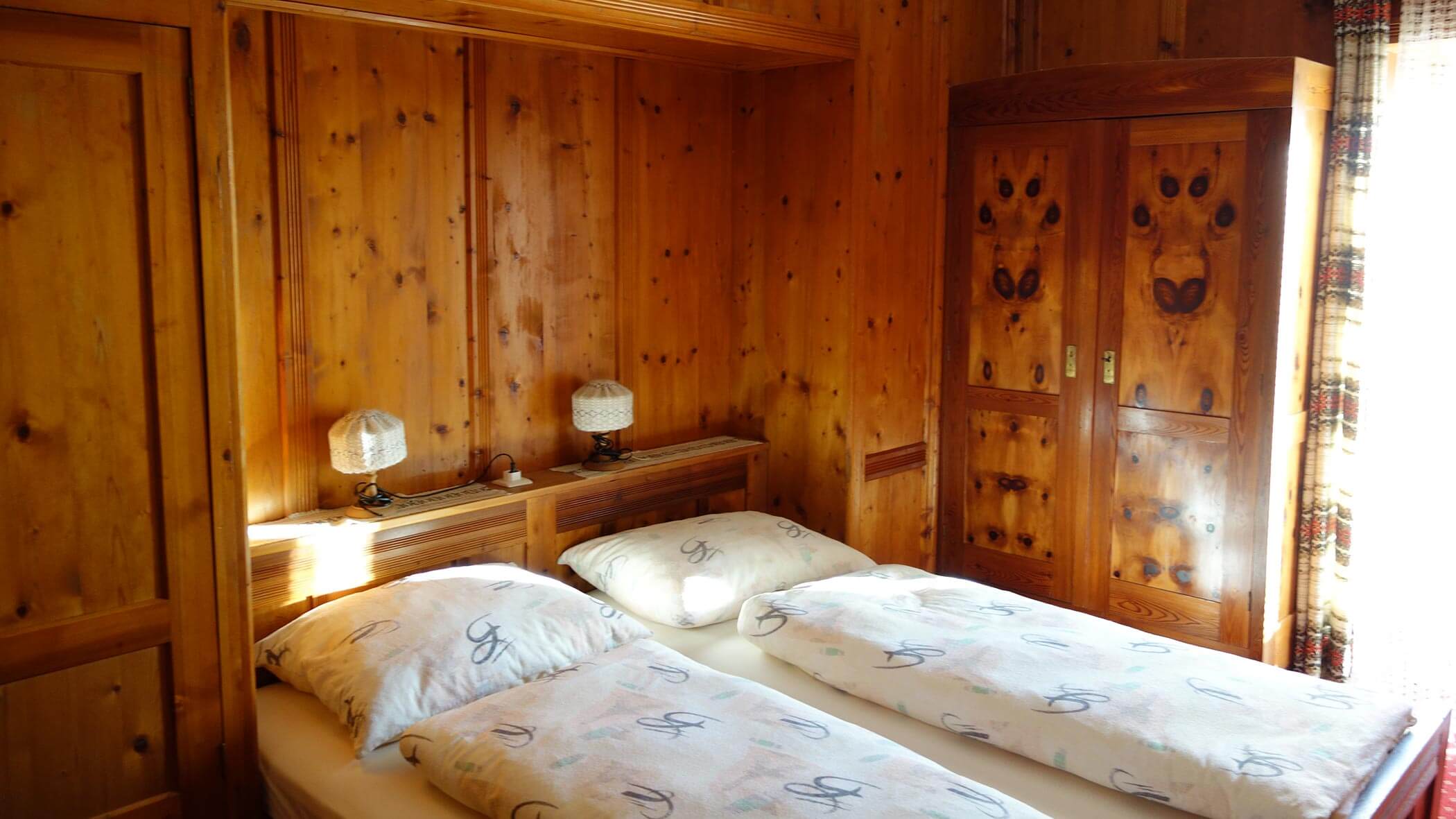 Gemütliches Holzschlafzimmer mit zwei Einzelbetten, gemusterten Bettdecken, Lampen und einem Kleiderschrank an einem sonnigen Fenster. - Garni Tschurtschenthaler