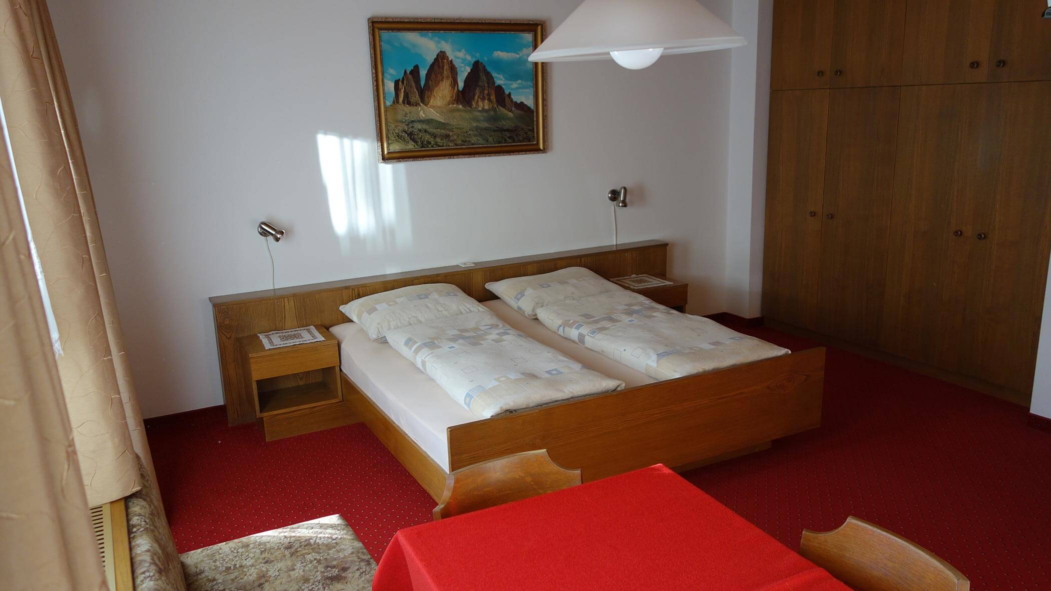 Ein ordentliches Zimmer mit zwei Betten, rotem Teppich, Holzmöbeln und einem Gemälde über dem Bett. - Garni Tschurtschenthaler