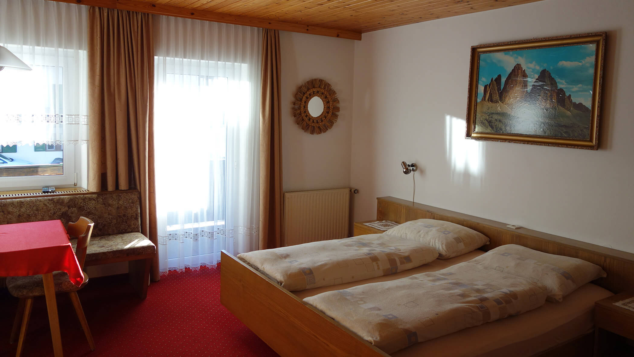 Gemütliches Zimmer mit Doppelbett, rotem Teppich, Essecke und einem Fenster mit durchsichtigen Vorhängen. - Garni Tschurtschenthaler