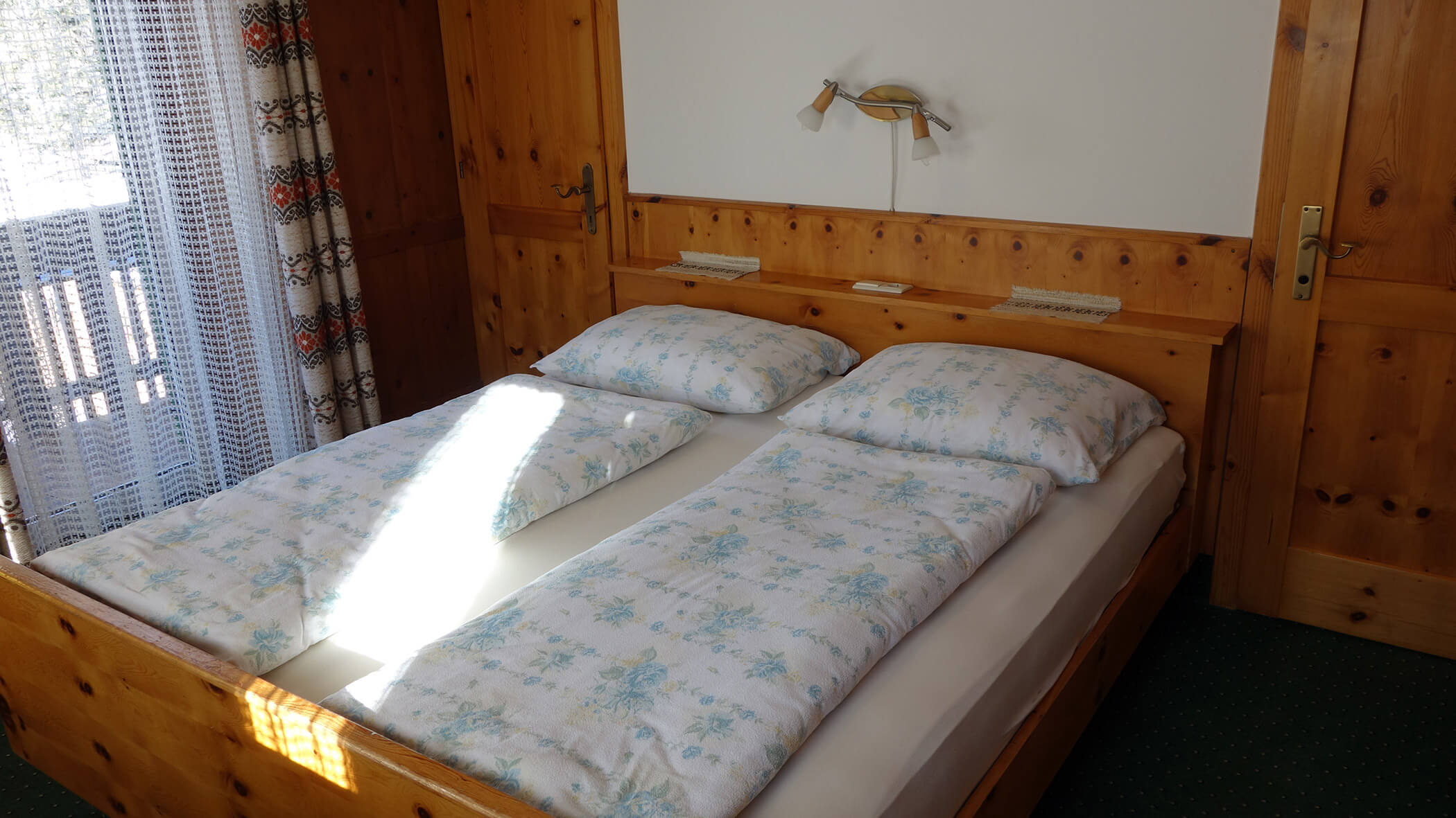 Ein Holzbett mit geblümtem Bettzeug neben einem Fenster mit Spitzenvorhängen und einfallendem Sonnenlicht. - Garni Tschurtschenthaler